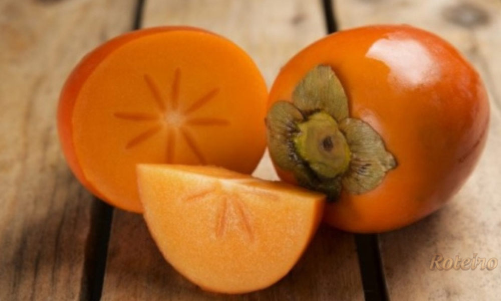 Caquis frescos sobre superfície branca: um inteiro, um cortado ao meio e um em quarto, mostrando a polpa laranja da fruta
