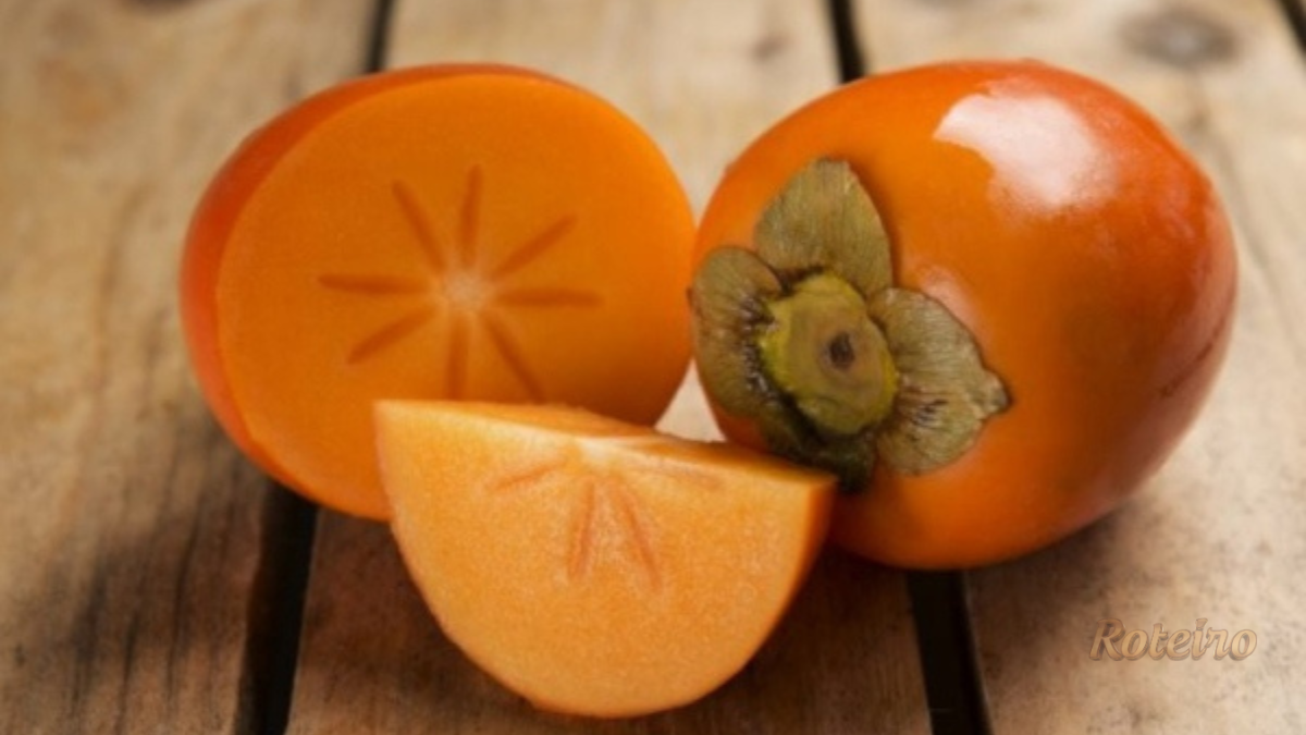 Caquis frescos sobre superfície branca: um inteiro, um cortado ao meio e um em quarto, mostrando a polpa laranja da fruta