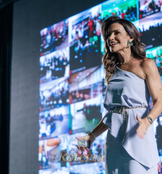 A escritora e palestrante Camila Vieira, uma mulher de cabelos castanhos claros e vestindo um vestido azul elegante, sorri e gesticula com entusiasmo em um grande evento. Ao fundo, um telão iluminado exibe vários fotos de vários eventos.