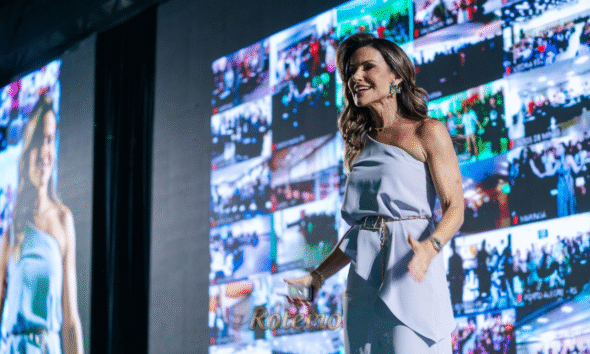 A escritora e palestrante Camila Vieira, uma mulher de cabelos castanhos claros e vestindo um vestido azul elegante, sorri e gesticula com entusiasmo em um grande evento. Ao fundo, um telão iluminado exibe vários fotos de vários eventos.
