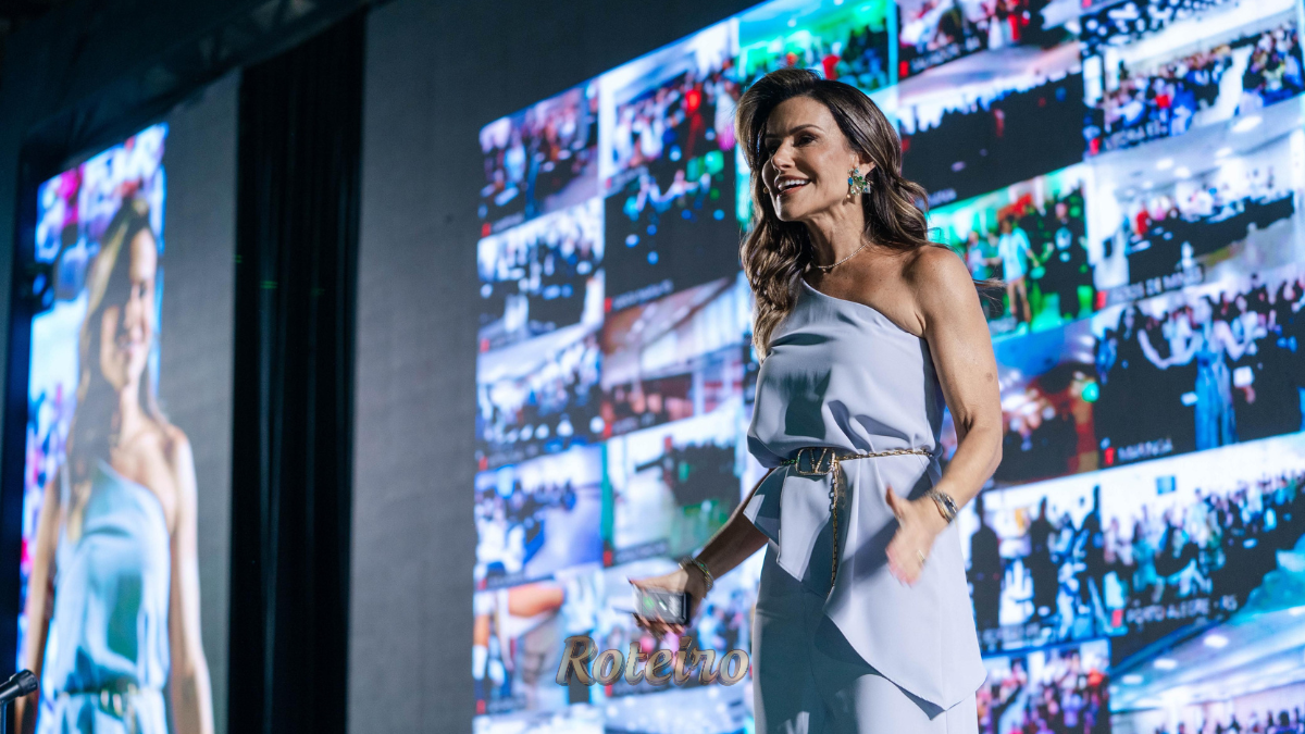 A escritora e palestrante Camila Vieira, uma mulher de cabelos castanhos claros e vestindo um vestido azul elegante, sorri e gesticula com entusiasmo em um grande evento. Ao fundo, um telão iluminado exibe vários fotos de vários eventos.