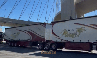 Comboio de caminhões modernos do Mirage Circus atravessando a Ponte Estaiada de São José dos Campos sob sol forte. Os veículos são grandes, coloridos e estampados com a marca do circo e a foto do ator Marcos Frota.