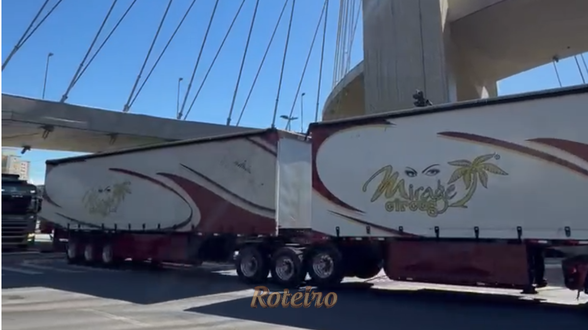 Comboio de caminhões modernos do Mirage Circus atravessando a Ponte Estaiada de São José dos Campos sob sol forte. Os veículos são grandes, coloridos e estampados com a marca do circo e a foto do ator Marcos Frota.