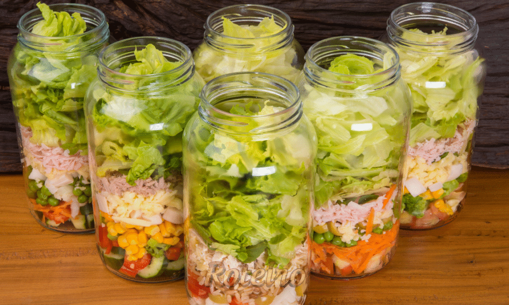 Vários potes de vidro alinhados com saladas coloridas em camadas, prontos para consumo. Exemplo de marmita saudável e econômica preparada para a semana.