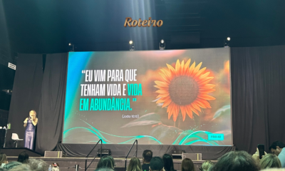 Palestrante Camila Viera em vestido azul no palco do espaço Roteiro, em São José dos Campos, durante apresentação ao vivo. Ao fundo, telão exibe girassol e versículo bíblico de João 10:10: "Eu vim para que tenham vida e vida em abundância.