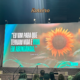 Palestrante Camila Viera em vestido azul no palco do espaço Roteiro, em São José dos Campos, durante apresentação ao vivo. Ao fundo, telão exibe girassol e versículo bíblico de João 10:10: "Eu vim para que tenham vida e vida em abundância.
