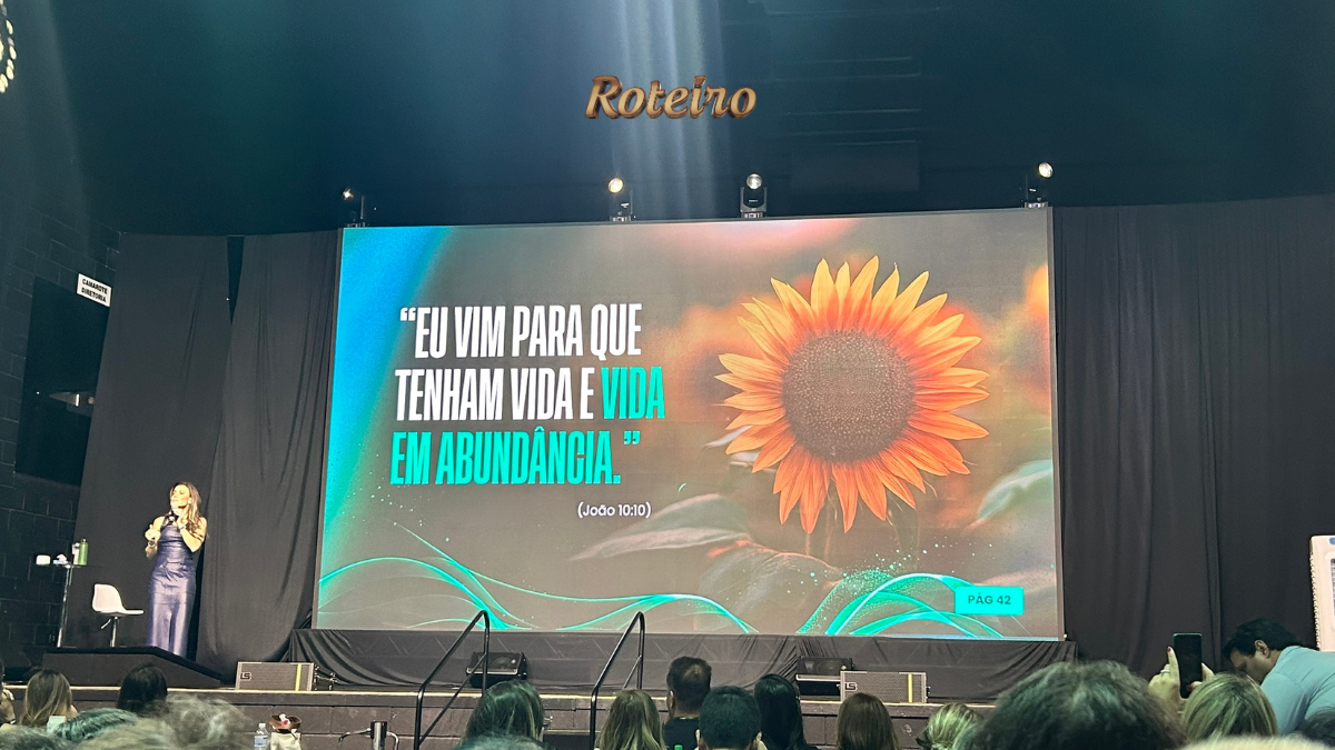 Palestrante Camila Viera em vestido azul no palco do espaço Roteiro, em São José dos Campos, durante apresentação ao vivo. Ao fundo, telão exibe girassol e versículo bíblico de João 10:10: "Eu vim para que tenham vida e vida em abundância.