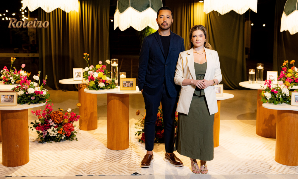 Casal de empreendedores Nadine Aldra e Rafael Yudi posam no espaço decorado do Aldras Experience em Jacareí, com mesas redondas de madeira, arranjos de flores coloridas, luminárias pendentes com franjas e quadros dispostos pelo ambiente ao fundo
