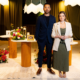 Casal de empreendedores Nadine Aldra e Rafael Yudi posam no espaço decorado do Aldras Experience em Jacareí, com mesas redondas de madeira, arranjos de flores coloridas, luminárias pendentes com franjas e quadros dispostos pelo ambiente ao fundo