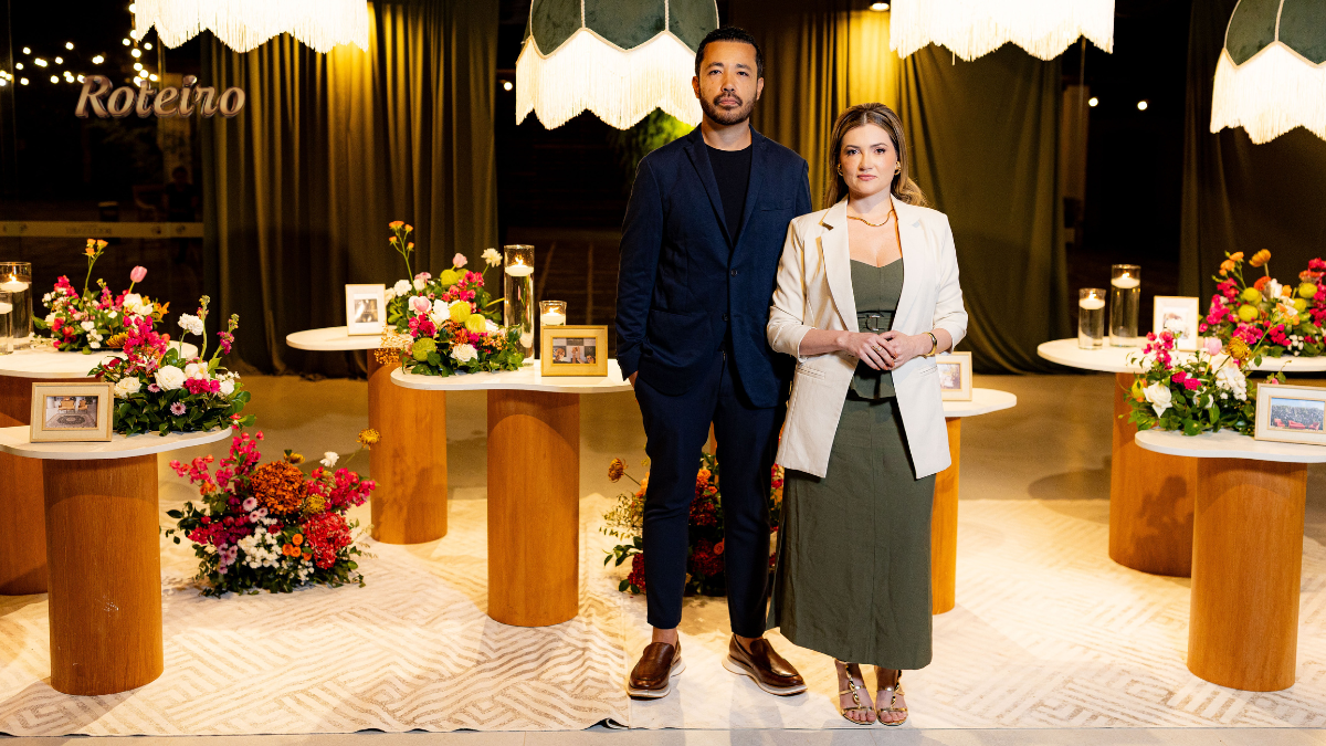 Casal de empreendedores Nadine Aldra e Rafael Yudi posam no espaço decorado do Aldras Experience em Jacareí, com mesas redondas de madeira, arranjos de flores coloridas, luminárias pendentes com franjas e quadros dispostos pelo ambiente ao fundo