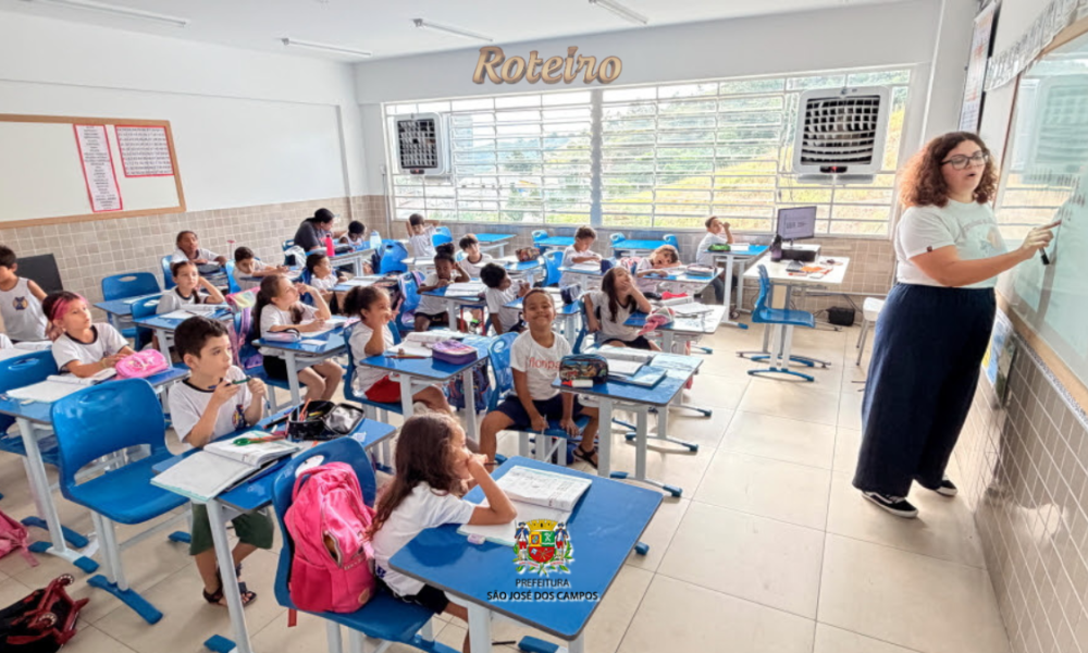 Sala de aula com crianças do ensino fundamental sentadas em carteiras azuis, uniformizadas com camiseta branca. Uma professora jovem, de cabelos cacheados e óculos, escreve no quadro branco à direita da imagem. O ambiente é bem iluminado, com janelas amplas e paredes de azulejo claro, em escola pública de São José dos Campos.