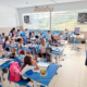 Sala de aula com crianças do ensino fundamental sentadas em carteiras azuis, uniformizadas com camiseta branca. Uma professora jovem, de cabelos cacheados e óculos, escreve no quadro branco à direita da imagem. O ambiente é bem iluminado, com janelas amplas e paredes de azulejo claro, em escola pública de São José dos Campos.