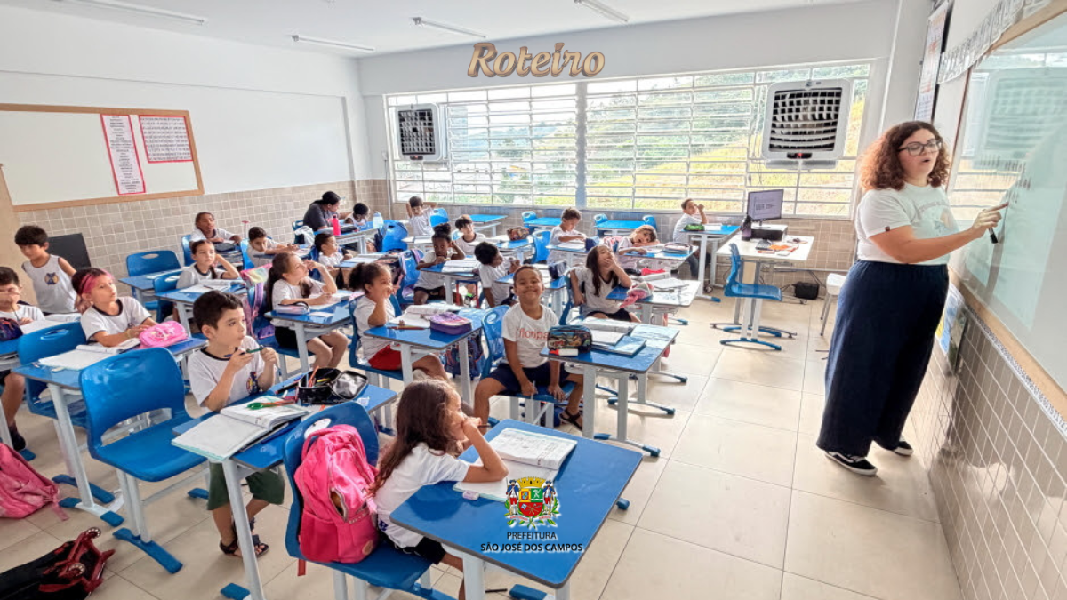 Sala de aula com crianças do ensino fundamental sentadas em carteiras azuis, uniformizadas com camiseta branca. Uma professora jovem, de cabelos cacheados e óculos, escreve no quadro branco à direita da imagem. O ambiente é bem iluminado, com janelas amplas e paredes de azulejo claro, em escola pública de São José dos Campos.
