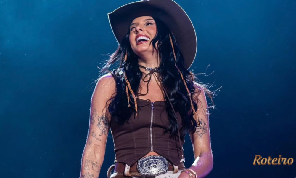 Ana Castela sorridente no palco, vestindo top marrom, chapéu country e cinto com fivela prateada, durante show com iluminação azul ao fundo. Foto: Divulgação