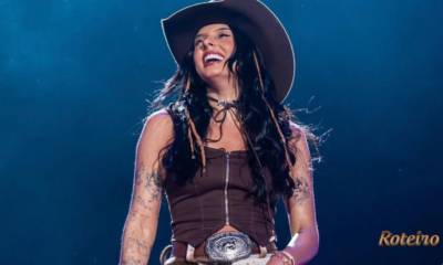 Ana Castela sorridente no palco, vestindo top marrom, chapéu country e cinto com fivela prateada, durante show com iluminação azul ao fundo. Foto: Divulgação