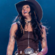 Ana Castela sorridente no palco, vestindo top marrom, chapéu country e cinto com fivela prateada, durante show com iluminação azul ao fundo. Foto: Divulgação