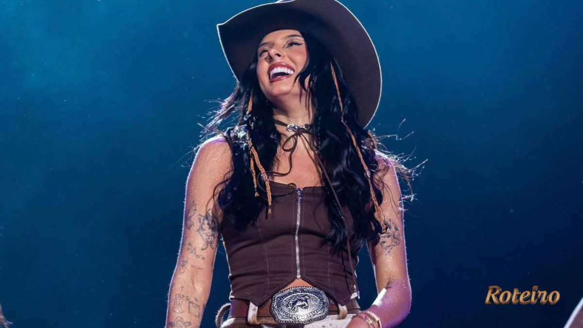 Ana Castela sorridente no palco, vestindo top marrom, chapéu country e cinto com fivela prateada, durante show com iluminação azul ao fundo. Foto: Divulgação