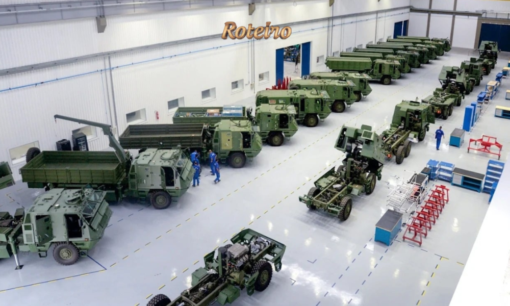 Vista interna de galpão industrial com veículos militares blindados e caminhões de combate na cor verde militar enfileirados, com funcionários com uniformes azuis trabalhando na linha de montagem. Foto: Divulgação/Avibras Aeroco