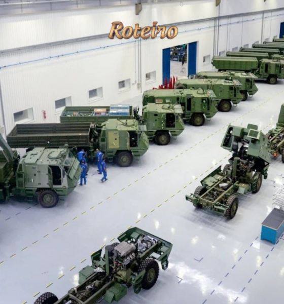 Vista interna de galpão industrial com veículos militares blindados e caminhões de combate na cor verde militar enfileirados, com funcionários com uniformes azuis trabalhando na linha de montagem. Foto: Divulgação/Avibras Aeroco
