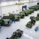 Vista interna de galpão industrial com veículos militares blindados e caminhões de combate na cor verde militar enfileirados, com funcionários com uniformes azuis trabalhando na linha de montagem. Foto: Divulgação/Avibras Aeroco