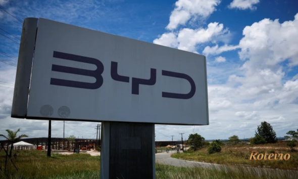 Totem com o logotipo da montadora BYD em área externa do complexo industrial de Camaçari, na Bahia, com estruturas da fábrica ao fundo e céu azul com nuvens. Marca registrada com letras em cinza escuro sobre painel metálico claro.