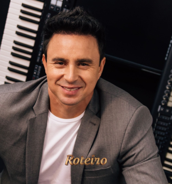 Allen Lima, músico integrante da Família Lima, sorridente em frente a dois teclados musicais, vestindo blazer cinza e camiseta branca em foto de divulgação do espetáculo Brasil em Teclas