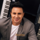 Allen Lima, músico integrante da Família Lima, sorridente em frente a dois teclados musicais, vestindo blazer cinza e camiseta branca em foto de divulgação do espetáculo Brasil em Teclas
