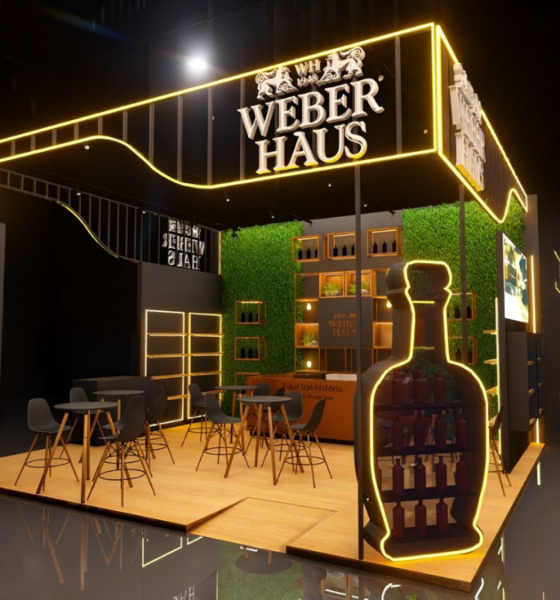 Estande da Weber Haus na APAS Show 2026 com iluminação dourada, parede de jardim vertical, prateleiras com garrafas de cachaça, mesas altas para degustação e silhueta luminosa de garrafa em destaque no primeiro plano