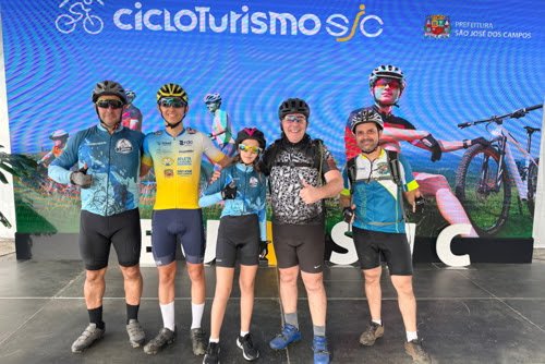 Grupo de cinco participantes posa em frente ao painel oficial do Cicloturismo SJC, com o brasão da Prefeitura de São José dos Campos ao lado da logo do programa, durante o evento de lançamento. Foto: Claudio Vieira/PMSJC