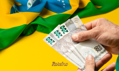 Pessoa segurando nota de dinheiro ao lado de decoração verde e amarela da Copa do Mundo 2026 — foto ilustrativa