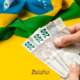 Pessoa segurando nota de dinheiro ao lado de decoração verde e amarela da Copa do Mundo 2026 — foto ilustrativa
