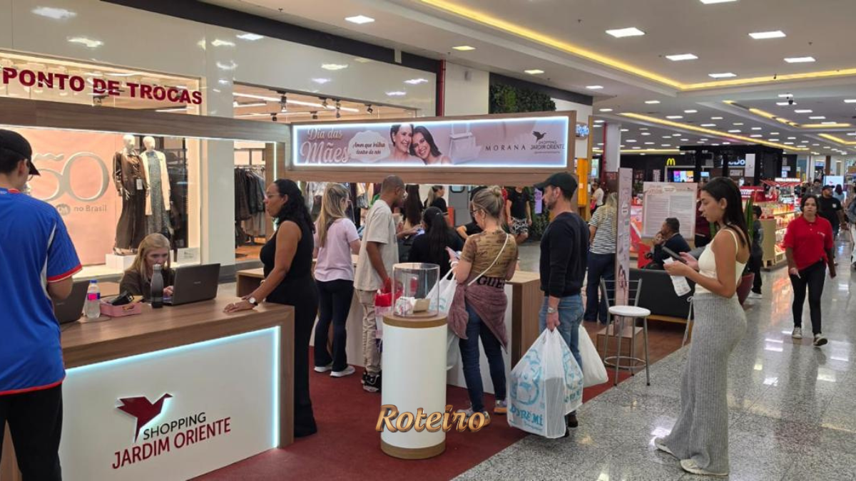 Clientes fazem fila no balcão "Ponto de Trocas" da campanha Dia das Mães do Shopping Jardim Oriente em São José dos Campos, com painel da Morana e movimento intenso no corredor do shopping ao fundo