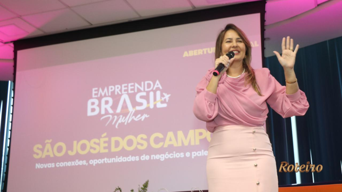 Fernanda Torres com blusa rosa segura microfone e gesticula durante abertura oficial do Empreenda Brasil Mulher em São José dos Campos, com projeção do evento ao fundo