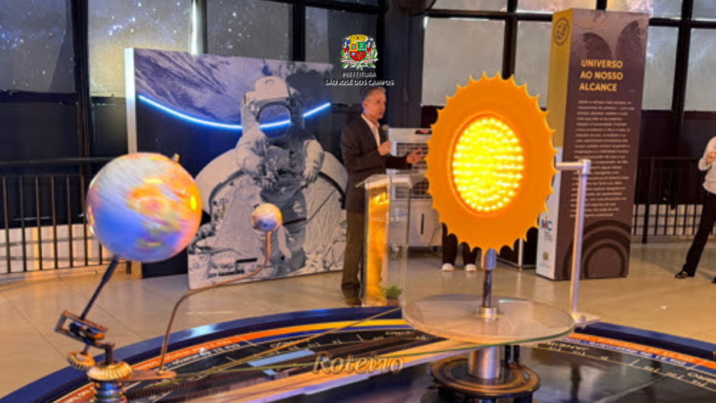 Palestrante discursa no Museu Interativo de Ciências de São José dos Campos diante de maquetes do sistema solar, com painel "Universo ao Nosso Alcance" ao fundo