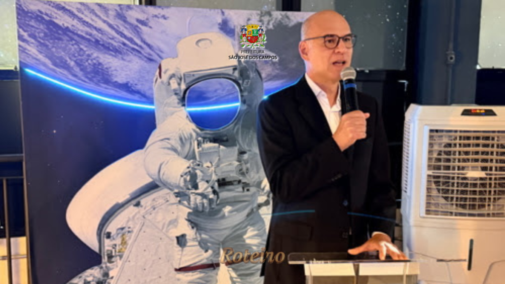 Homem de terno preto discursa ao microfone em púlpito durante inauguração da exposição Raios! Mensageiros do Cosmos no MIC, com painel de astronauta ao fundo