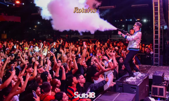 Artista no palco da festa Sub-18 Teen dispara canhão de fumaça colorida sobre multidão de adolescentes animados com os braços erguidos, durante evento noturno ao ar livre em São José dos Campos. Logo Sub-18 Teen visível na parte inferior da imagem. Foto: Divulgação