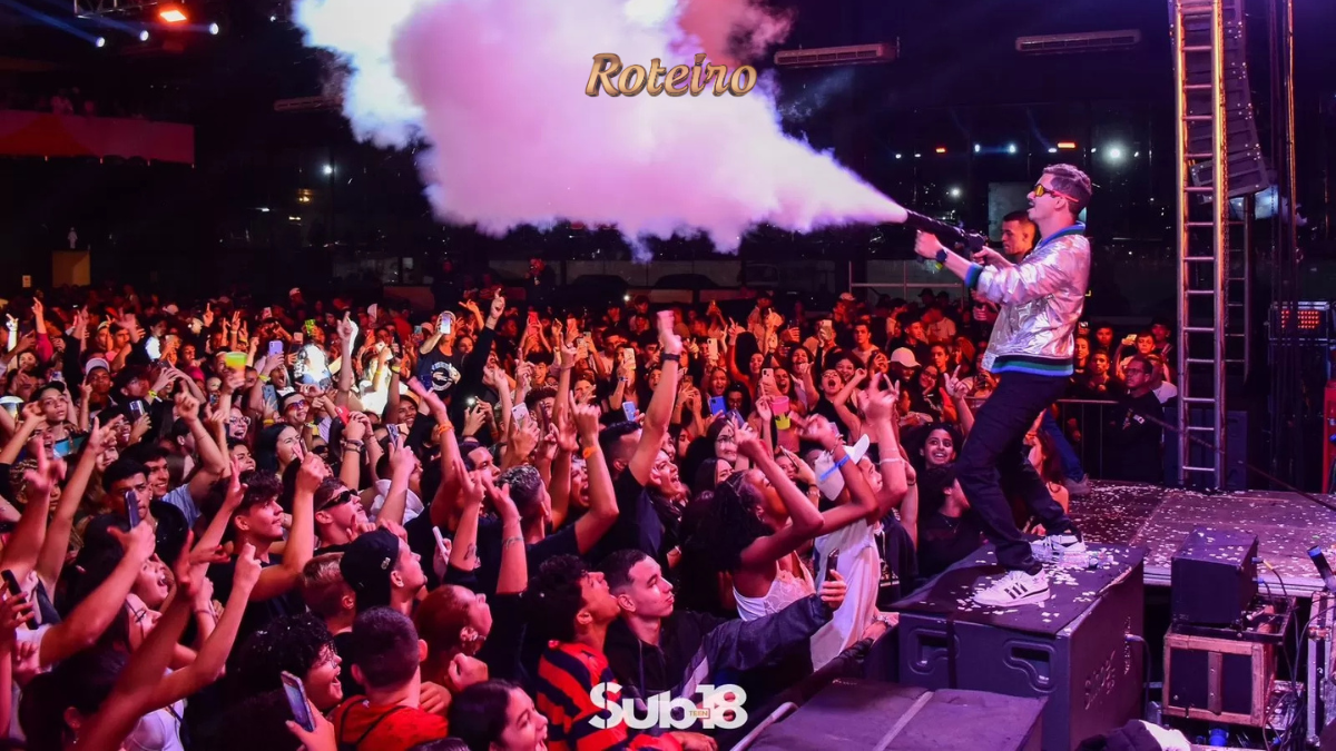 Artista no palco da festa Sub-18 Teen dispara canhão de fumaça colorida sobre multidão de adolescentes animados com os braços erguidos, durante evento noturno ao ar livre em São José dos Campos. Logo Sub-18 Teen visível na parte inferior da imagem. Foto: Divulgação
