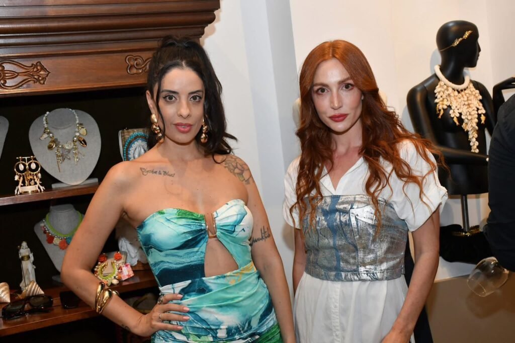 Duas convidadas elegantemente vestidas posam em frente à estante de joias e acessórios da Gregga Store durante o evento de aniversário em SJC.