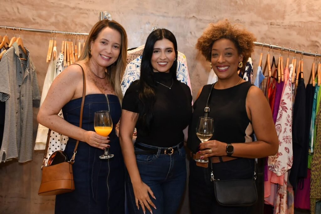 Trio de convidadas sorridentes com looks variados e taças de drinques posam em frente ao arara de roupas coloridas da Gregga Store durante evento de aniversário em SJC.