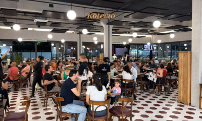 Salão lotado do Bar Coronel durante evento noturno, com dezenas de clientes sentados em mesas redondas de madeira, bebendo e conversando. O ambiente tem piso de ladrilho hidráulico branco e bordô, luminárias esféricas pendentes e grandes janelas com vista para a rua iluminada. Ao fundo, telão com a frase "O nosso é fazer isso acontecer."