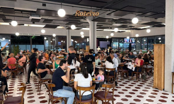 Salão lotado do Bar Coronel durante evento noturno, com dezenas de clientes sentados em mesas redondas de madeira, bebendo e conversando. O ambiente tem piso de ladrilho hidráulico branco e bordô, luminárias esféricas pendentes e grandes janelas com vista para a rua iluminada. Ao fundo, telão com a frase "O nosso é fazer isso acontecer."