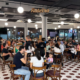 Salão lotado do Bar Coronel durante evento noturno, com dezenas de clientes sentados em mesas redondas de madeira, bebendo e conversando. O ambiente tem piso de ladrilho hidráulico branco e bordô, luminárias esféricas pendentes e grandes janelas com vista para a rua iluminada. Ao fundo, telão com a frase "O nosso é fazer isso acontecer."