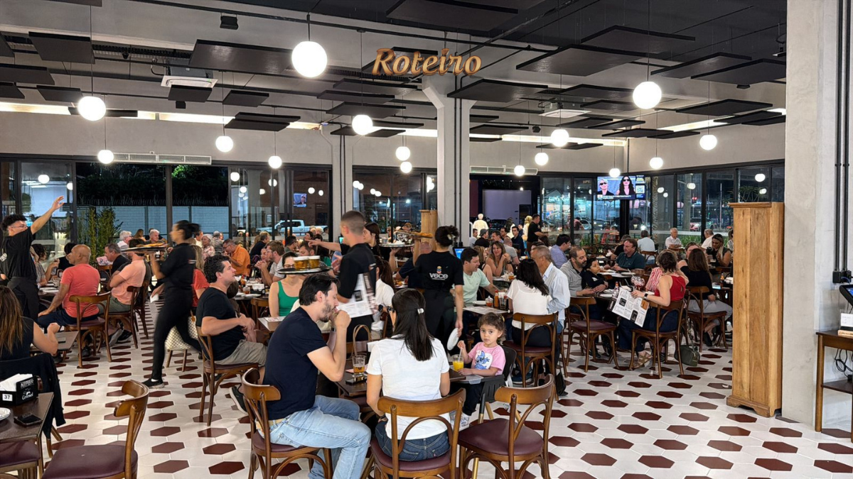 Salão lotado do Bar Coronel durante evento noturno, com dezenas de clientes sentados em mesas redondas de madeira, bebendo e conversando. O ambiente tem piso de ladrilho hidráulico branco e bordô, luminárias esféricas pendentes e grandes janelas com vista para a rua iluminada. Ao fundo, telão com a frase "O nosso é fazer isso acontecer."