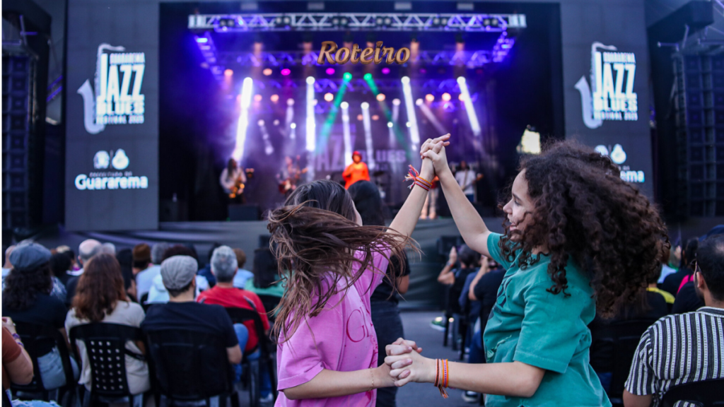 Duas meninas dançam em frente ao palco do Guararema Jazz & Blues Festival com público e iluminação ao fundo