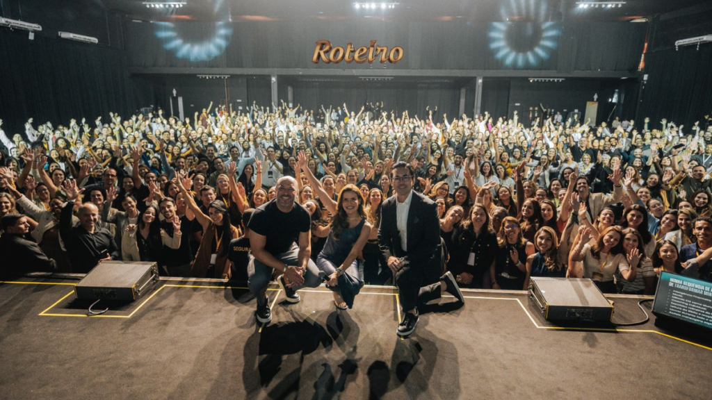 Foto panorâmica do palco do Espaço Sunset, em São José dos Campos, durante o evento da Camila Viera “Viva a Sua Real Identidade” com plateia lotada de braços erguidos. Três palestrantes posam sentados na borda do palco comemorando o evento