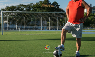 Jogador veterano com camiseta regata vermelha e short branco posa com o pé sobre uma bola de futebol em campo gramado de São José dos Campos, com trave ao fundo, em imagem de divulgação do Campeonato Joseense 50+ e 60+ da Prefeitura de SJC. Foto: PMSJC