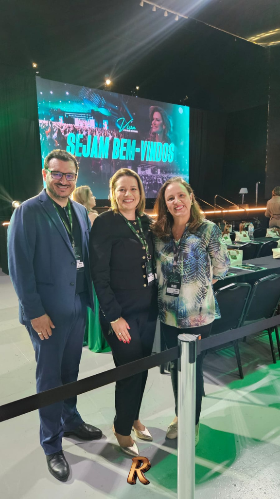 Johnis, Liliane Santos e Patricia Morello posam juntos no evento Viva, em São José dos Campos. Os três sorriem em frente ao telão com a mensagem "Sejam Bem-Vindos" e a logo do evento. Johnis veste terno azul com crachá; Liliane Santos, blazer preto com crachá; Patricia Morello, blusa estampada com crachá