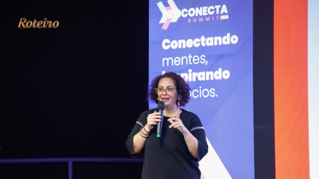Palestrante do ProConecta Summit no palco, mulher de blusa preta e óculos de armação colorida, cabelos cacheados, segurando microfone com expressão de engajamento. Ao fundo, painel do evento com os dizeres "Conectando mentes, inspirando negócios."