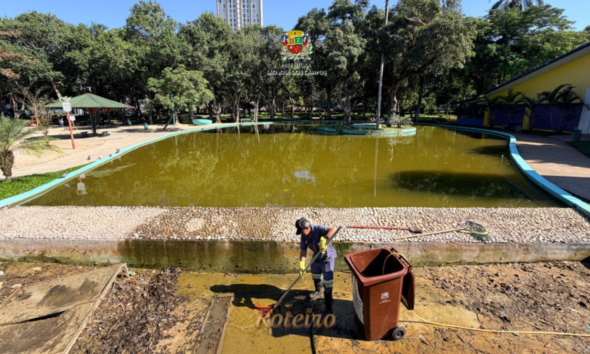Funcionário da Prefeitura de São José dos Campos com uniforme e luvas amarelas realiza limpeza do lago principal do Parque Santos Dumont, com o espelho d'água esverdeado ao fundo e lixeira marrom ao lado, durante manutenção periódica. Foto: Claudio Vieira/PMSJC