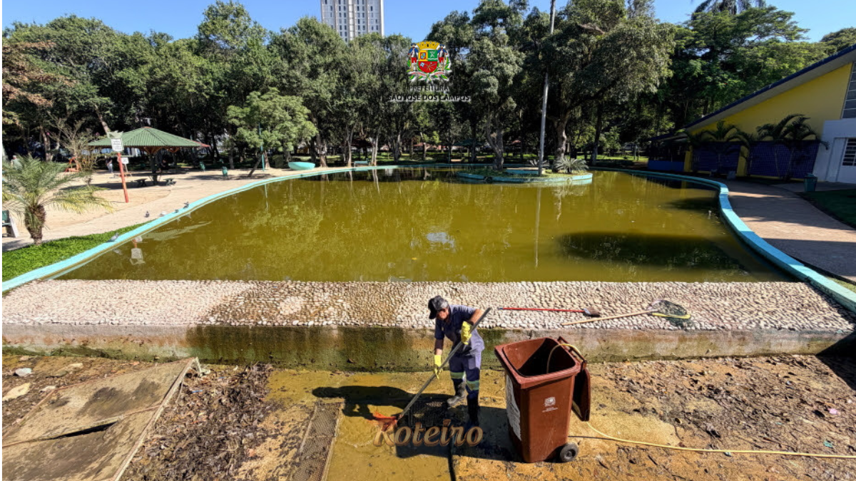 Funcionário da Prefeitura de São José dos Campos com uniforme e luvas amarelas realiza limpeza do lago principal do Parque Santos Dumont, com o espelho d'água esverdeado ao fundo e lixeira marrom ao lado, durante manutenção periódica. Foto: Claudio Vieira/PMSJC